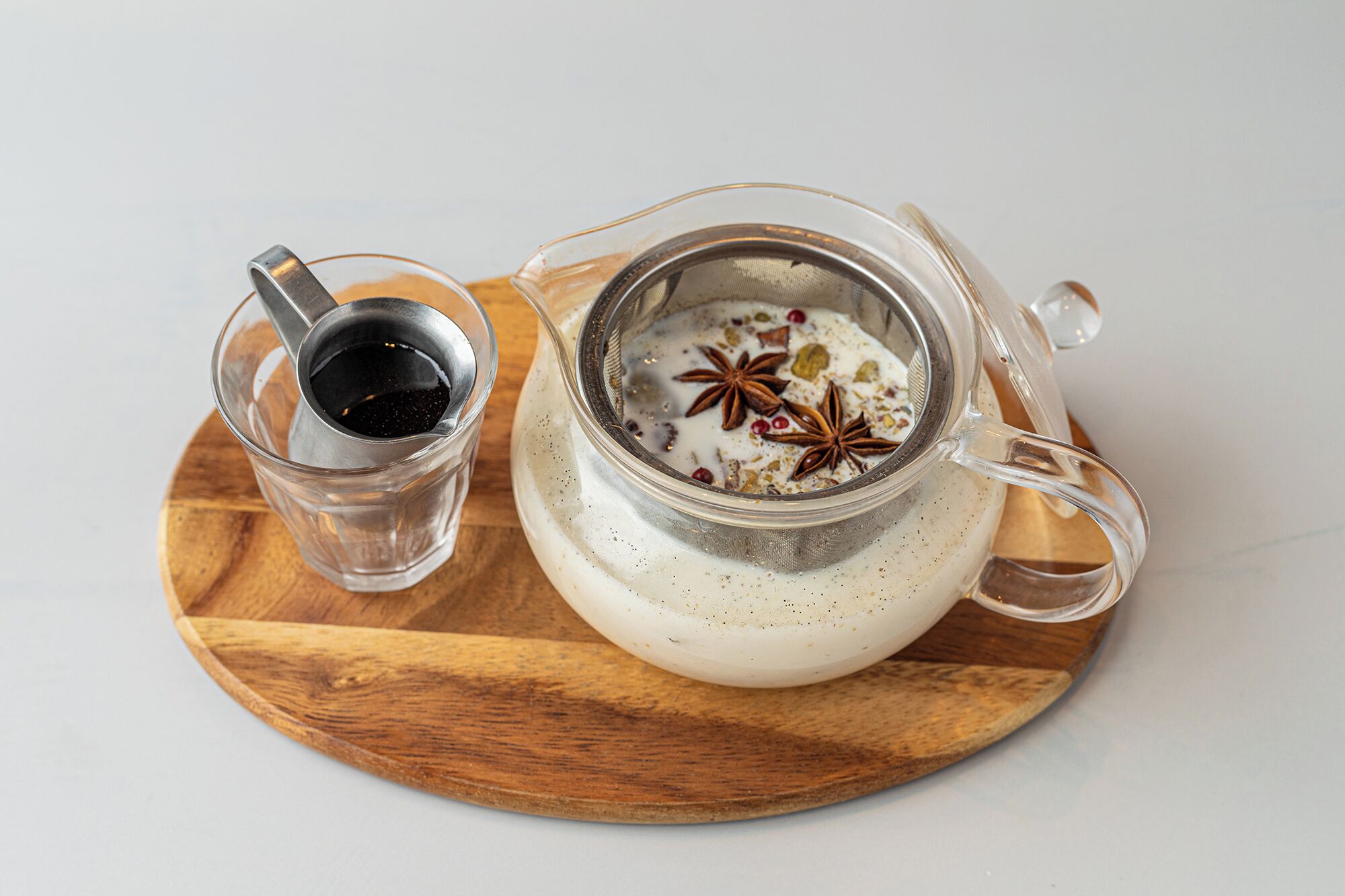 A clear glass teapot with a metal infuser and a glass cup with a metal spoon on a wooden board.