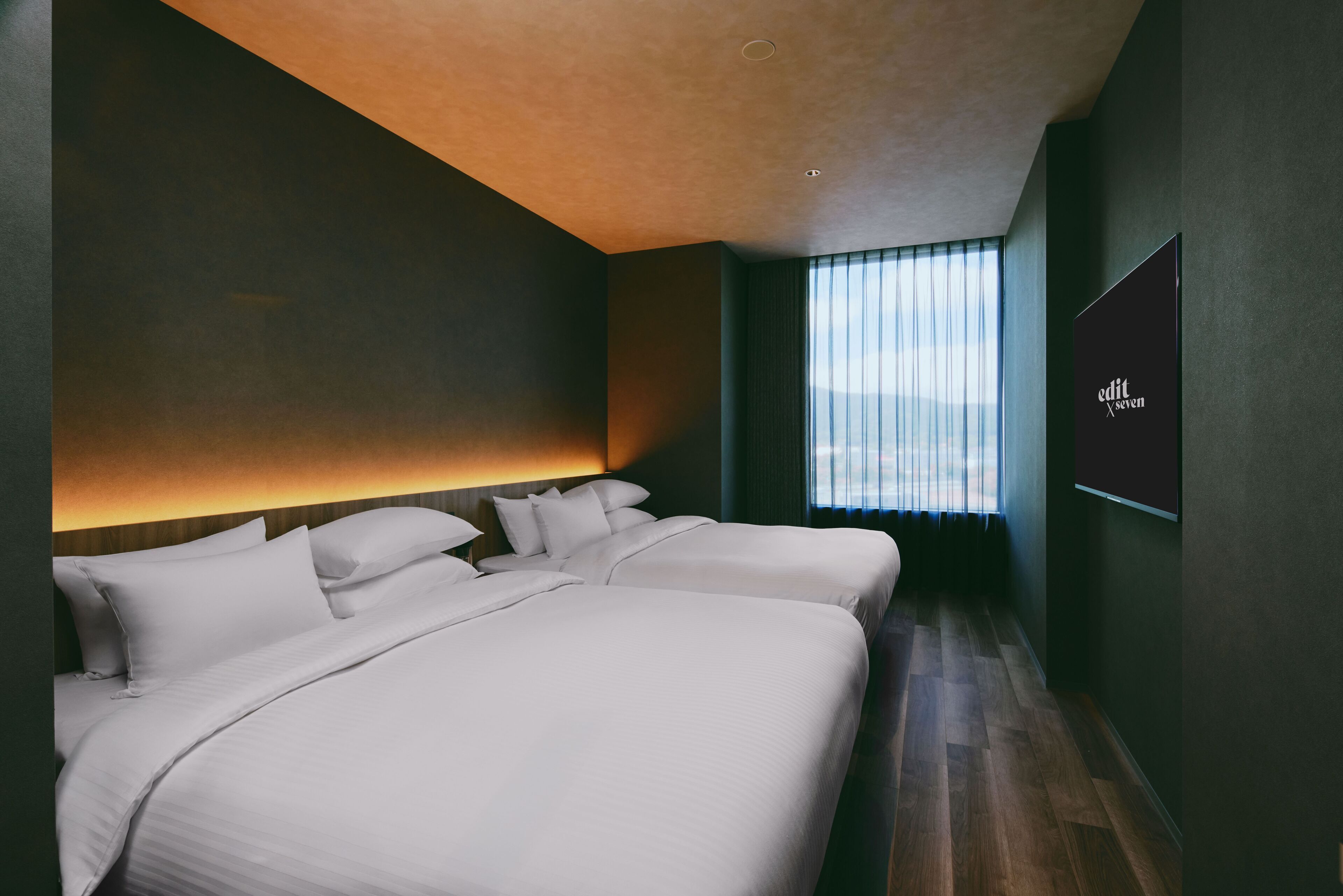 A modern hotel room with two beds, a large window with blinds, and a wall-mounted TV.