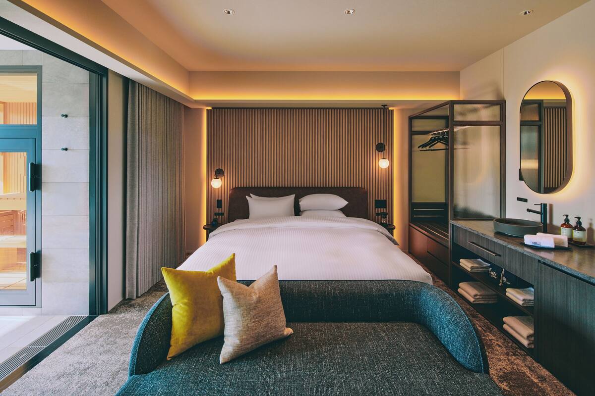 Ein modernes Hotelzimmer mit einem großen Bett, einer Sitzecke mit Kissen, einem Schminktisch mit Spiegel und Blick durch eine Glastür nach außen.