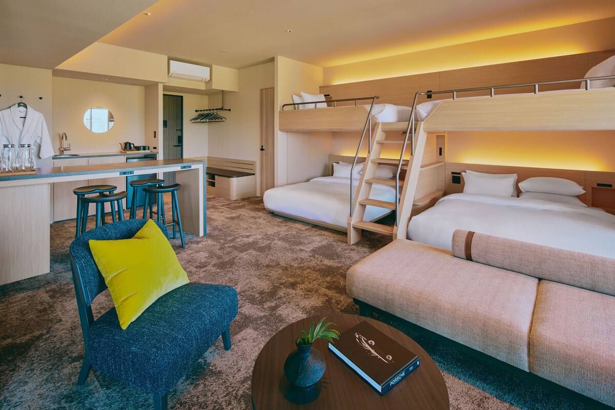Ein Hotelzimmer mit einem Etagenbett, einem Schreibtisch mit Hocker, einem Sessel mit gelbem Kissen und einem kleinen Tisch mit Pflanze und Buch.