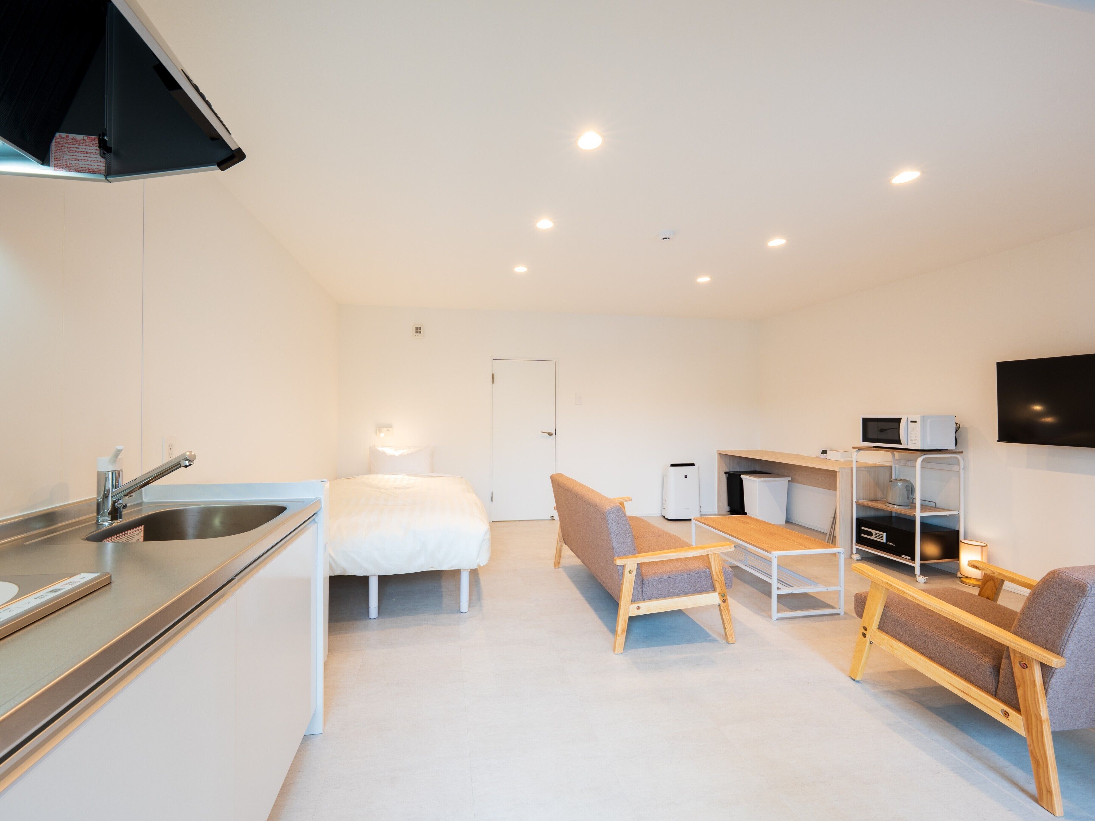 A modern, well-lit room with a kitchenette, a bed, a seating area with a table and chairs, and a flat-screen TV mounted on the wall.
