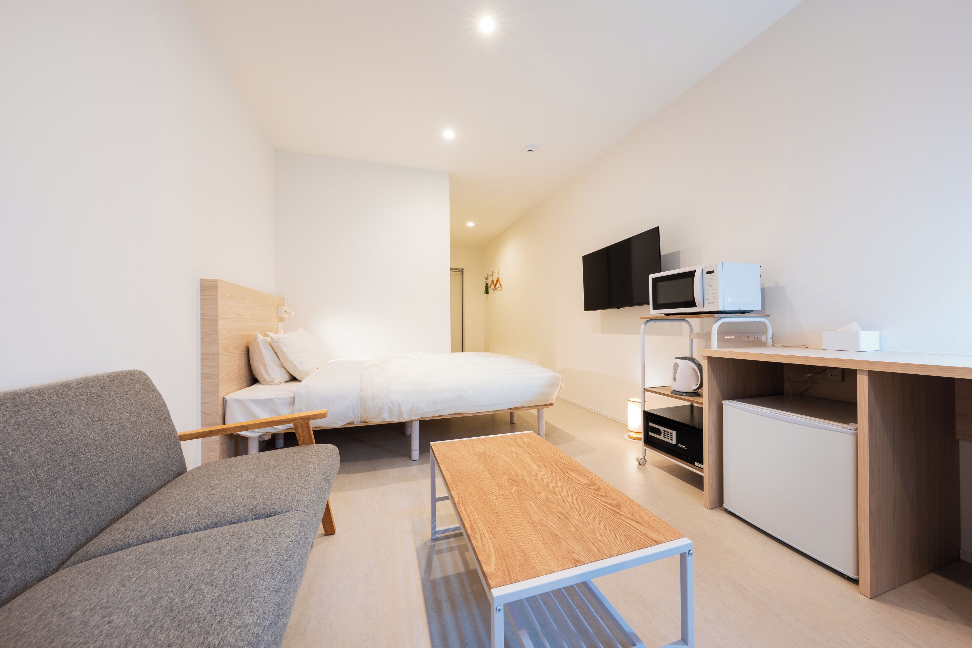 A compact hotel room with a bed, a sofa, a small table, a desk with a computer, a microwave, and a TV.