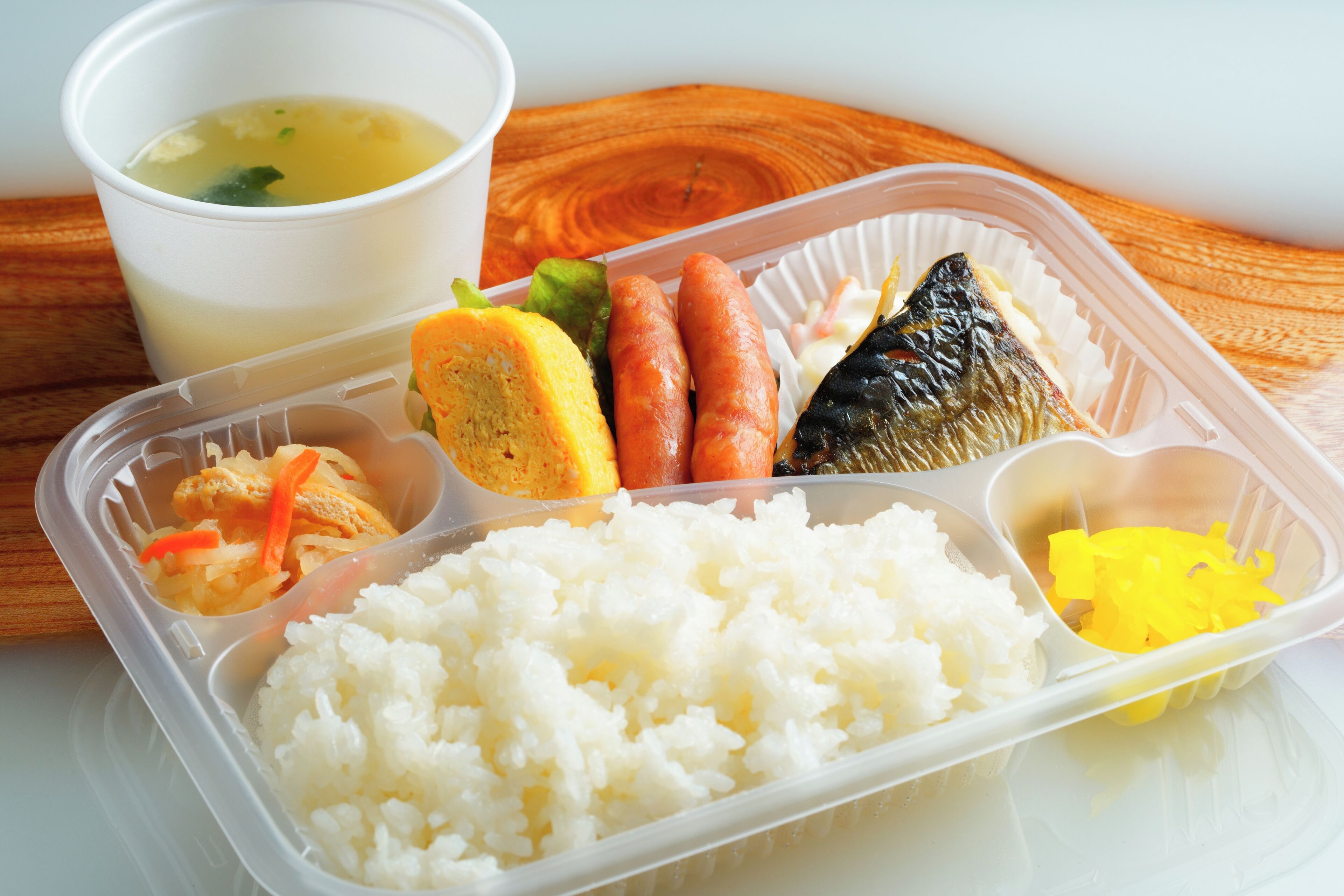 A meal in a plastic container with white rice, fish, carrots, and a soup cup.
