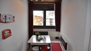 Una habitación pequeña con una mesa blanca, taburetes rojos, un banco rojo, una ventana con cortinas y una planta sobre la mesa.