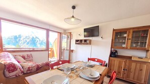 A dining area with a table set for four, wooden cabinets, a television on the wall, and a view of mountains through a large window. - Ski-In Stay in Les Deux Alpes (Les Deux Alpes)