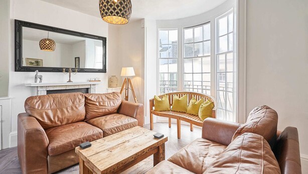 A living room with a brown leather sofa, a wooden coffee table, and a yellow armchair. There is a fireplace with a white mantel, a large mirror, and a hanging light fixture. - Host Stay Bedford Street (Brighton)
