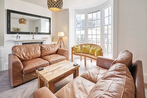 A living room with a brown leather sofa, a wooden coffee table, and a yellow armchair. There is a fireplace with a white mantel, a large mirror, and a hanging light fixture. - Host Stay Bedford Street (Brighton)
