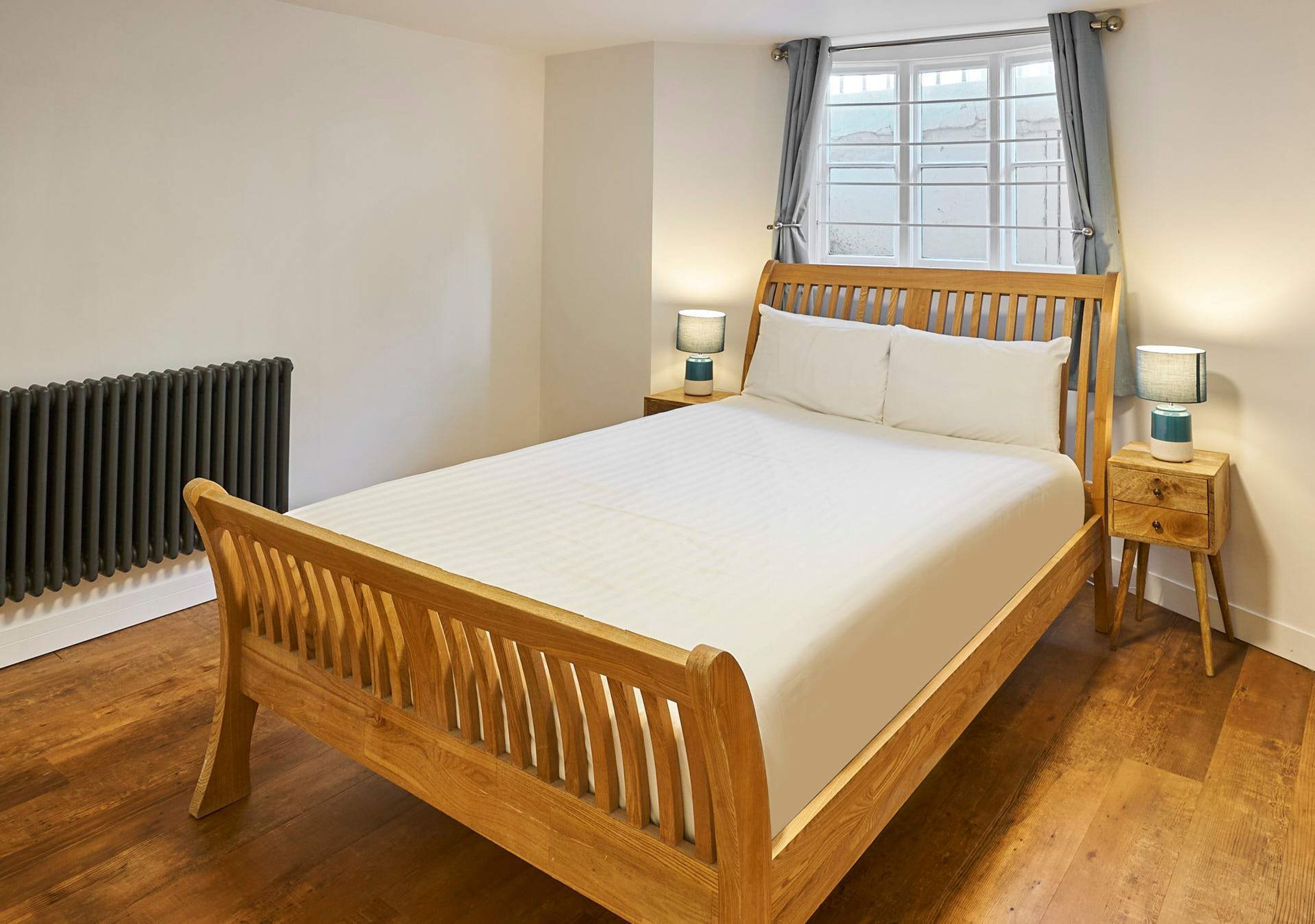 Un lit en bois avec un matelas blanc, dans une pièce dont le sol est recouvert de planches de bois, avec un radiateur et deux tables de chevet équipées de lampes.