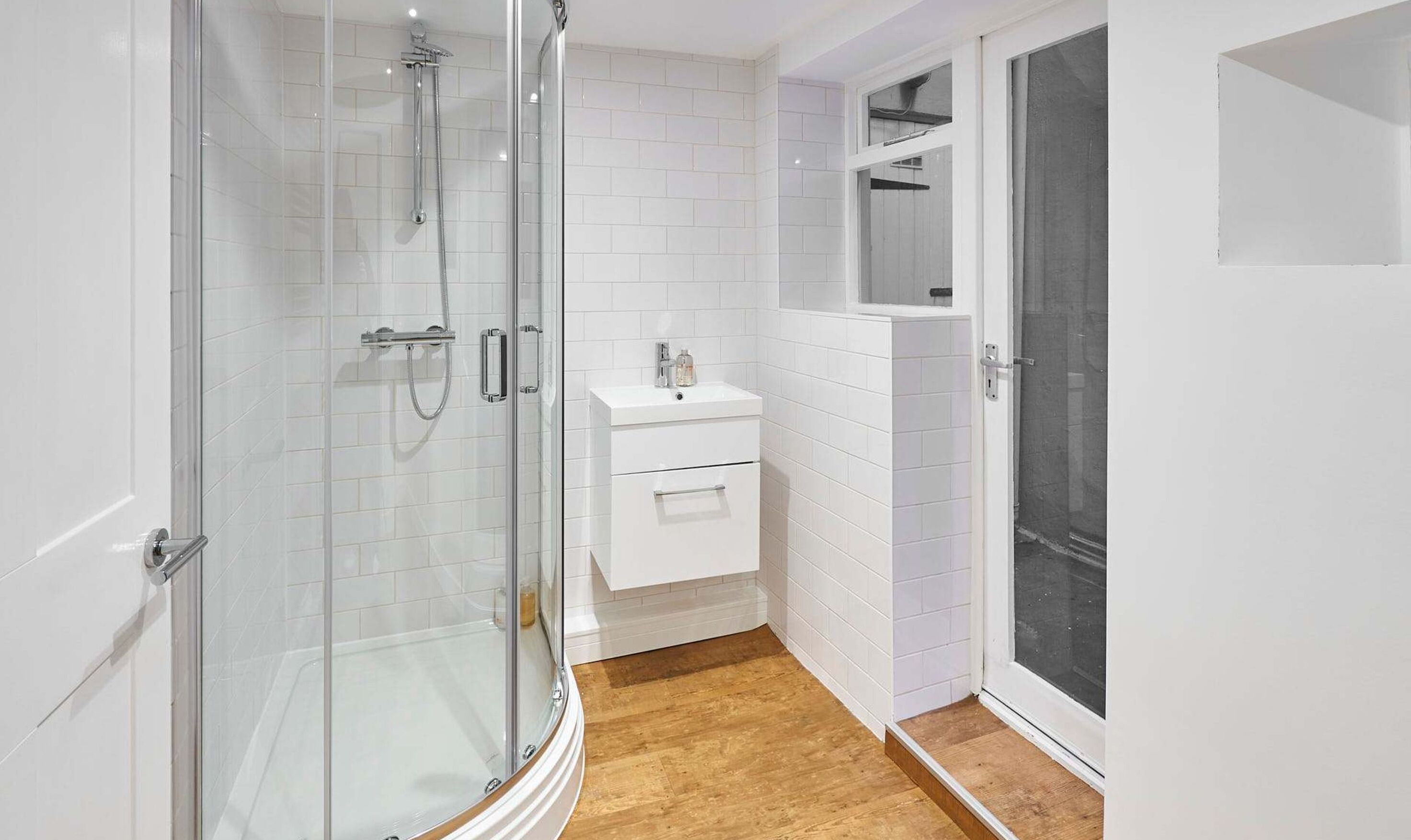 A modern bathroom with a glass-enclosed shower, a white vanity with a mirror, and a wooden floor.