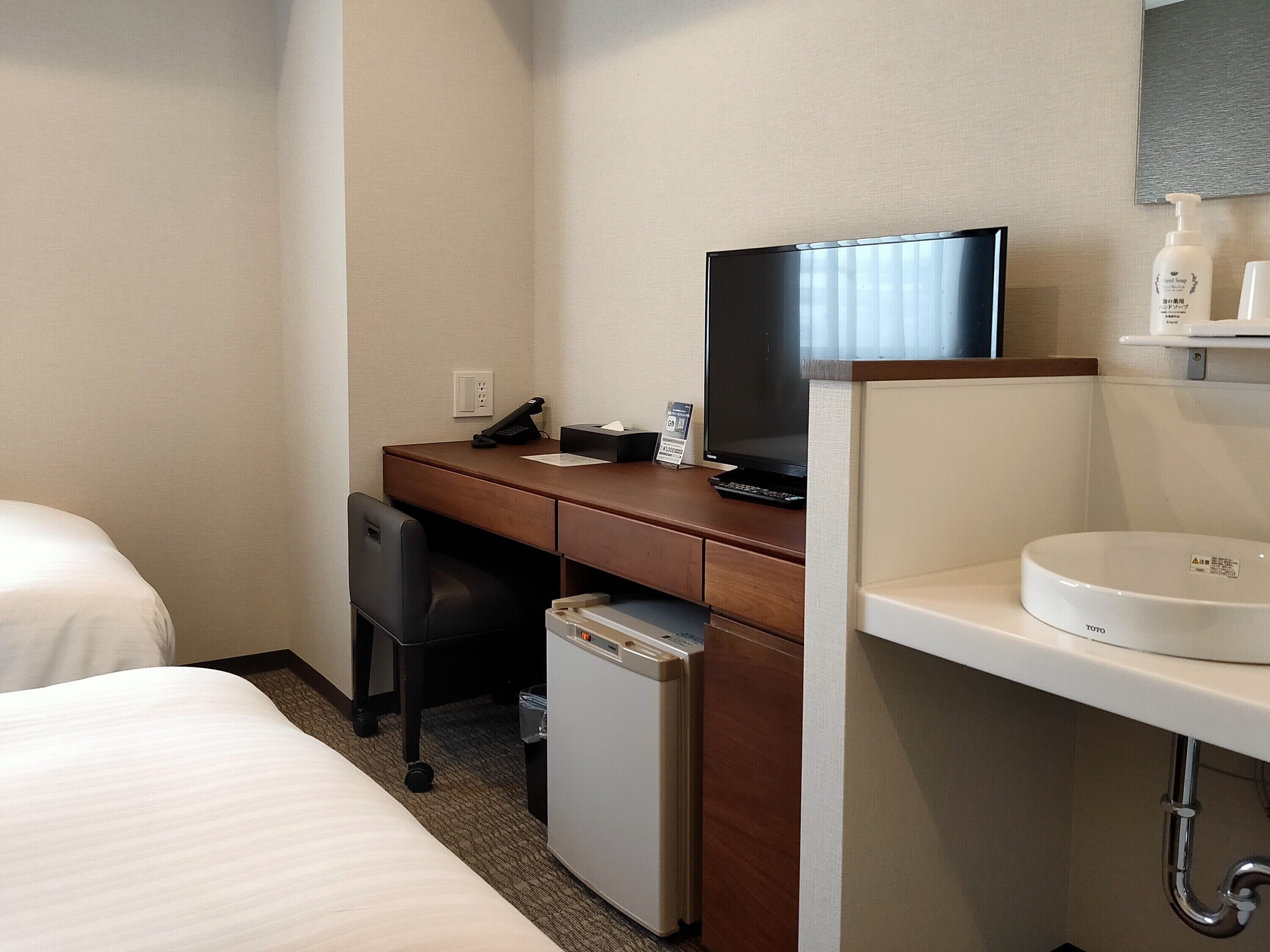 Een hotelkamer met een bed, een bureau met een computer, een televisie, een kleine koelkast en een wastafel.