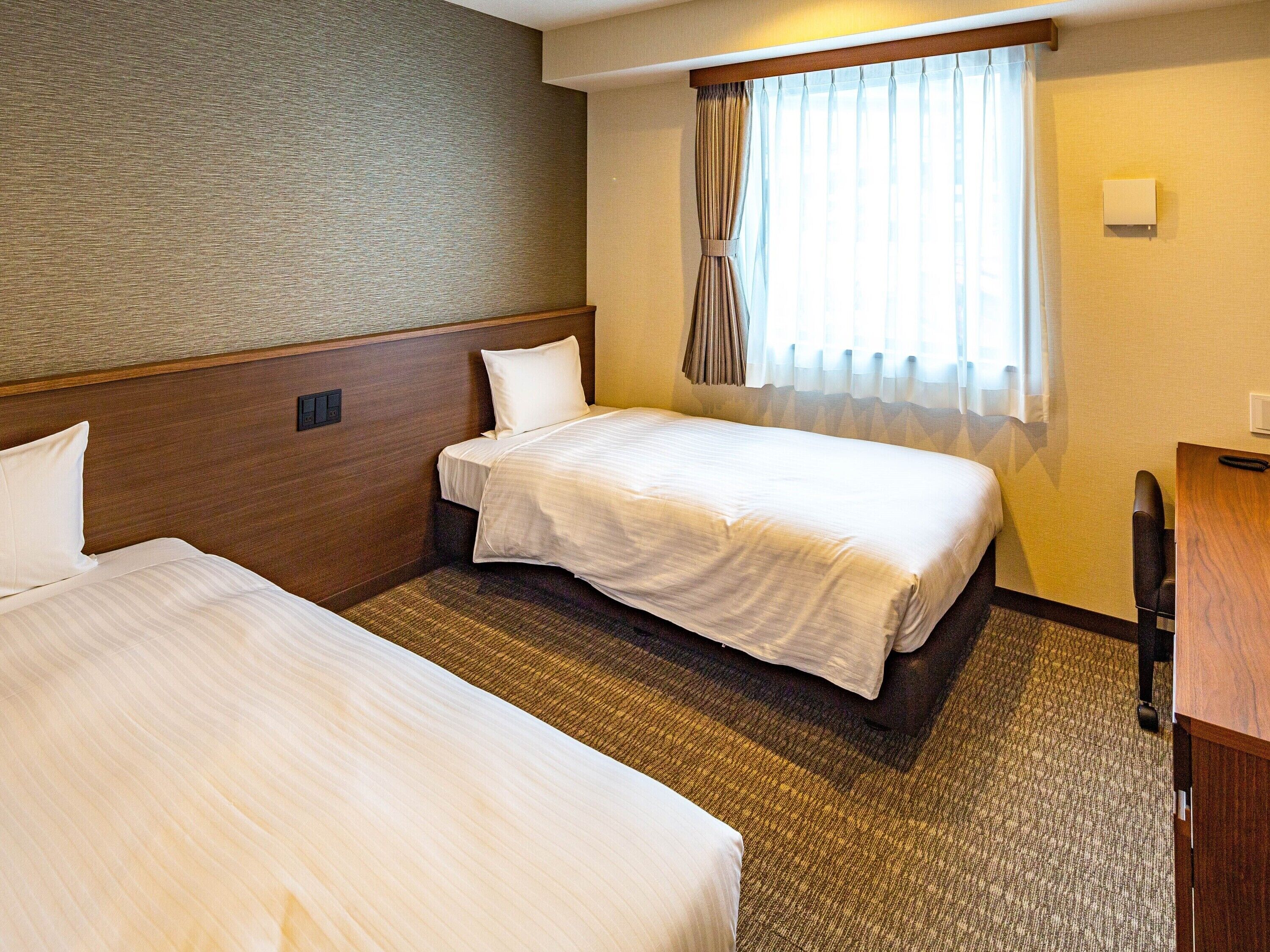 A hotel room with two beds, a wooden headboard, a carpeted floor, a window with curtains, and a desk with a chair.