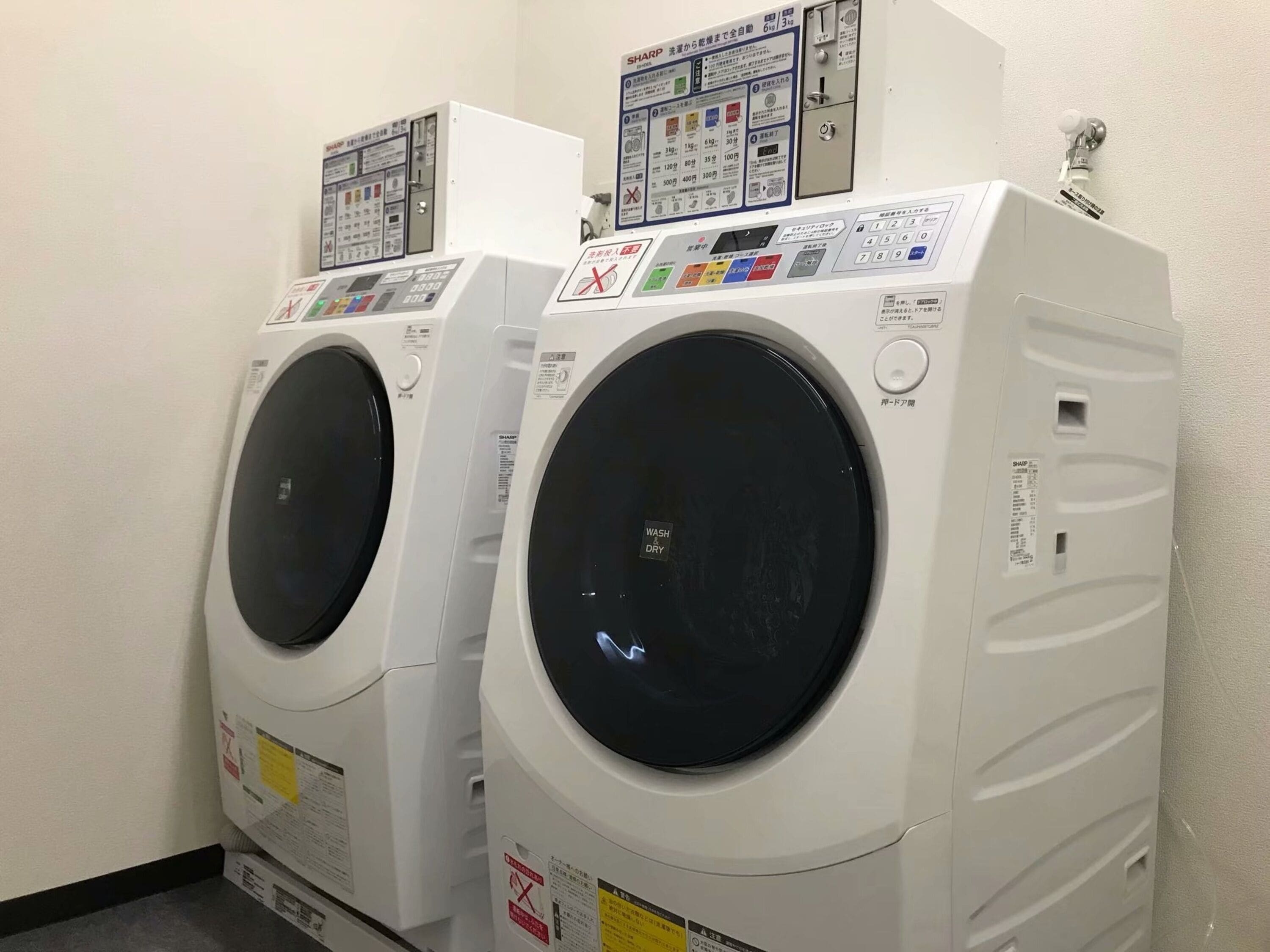 A row of white front-loading washing machines in a laundry room with control panels and safety instructions.