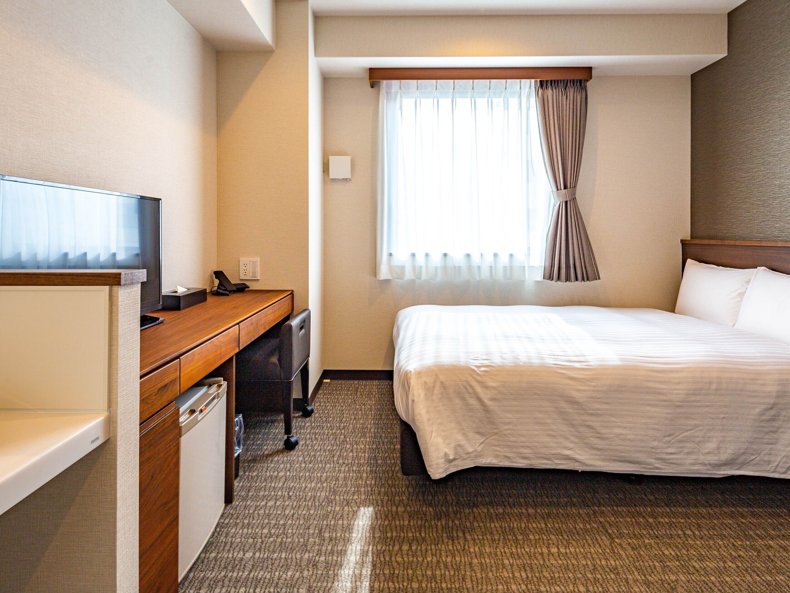 Une chambre d’hôtel comprenant un lit, un bureau avec une télévision, un téléphone et une fenêtre avec des rideaux.