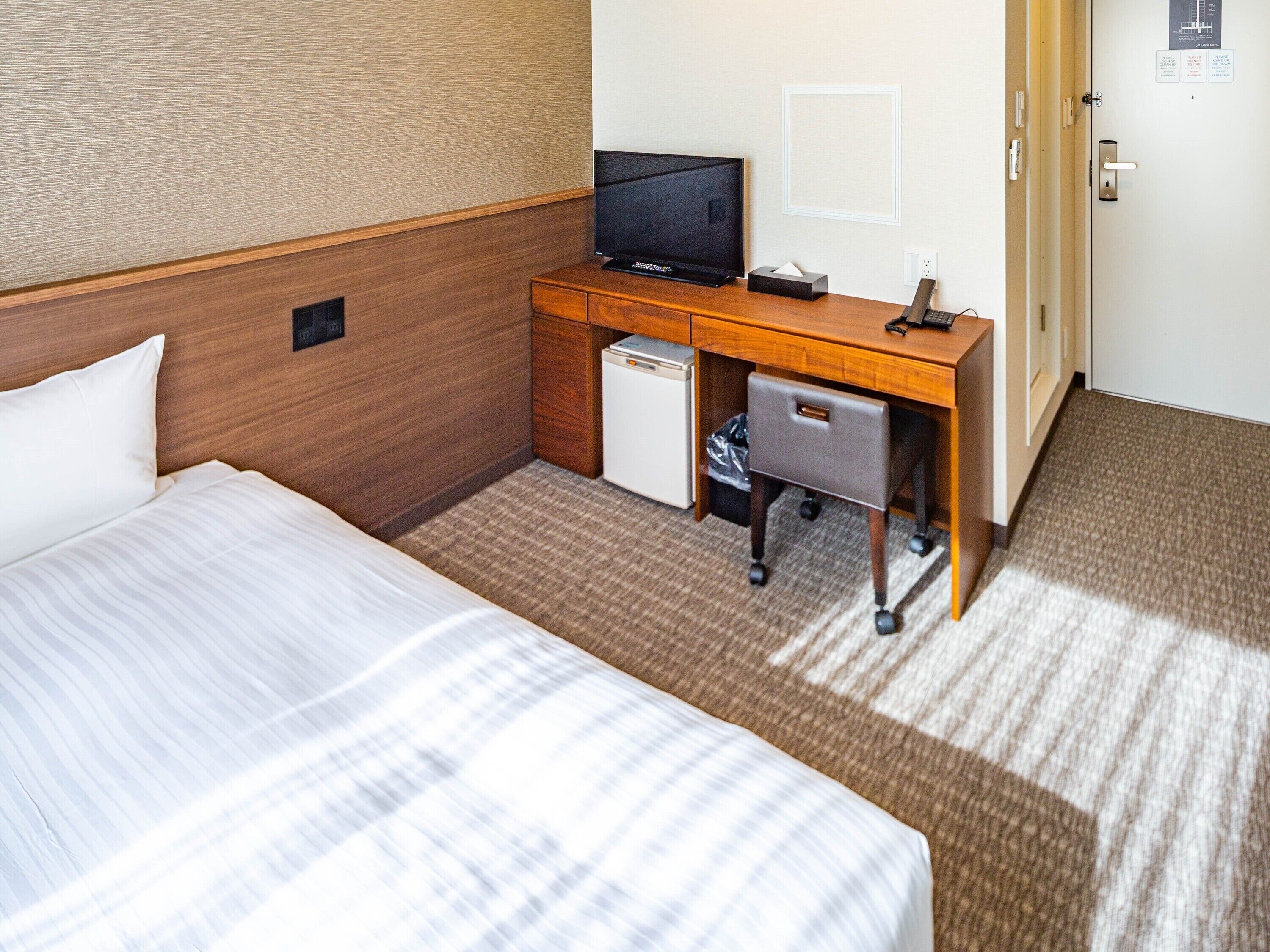 A hotel room with a bed, a desk with a television, a small refrigerator, and a trash bin.
