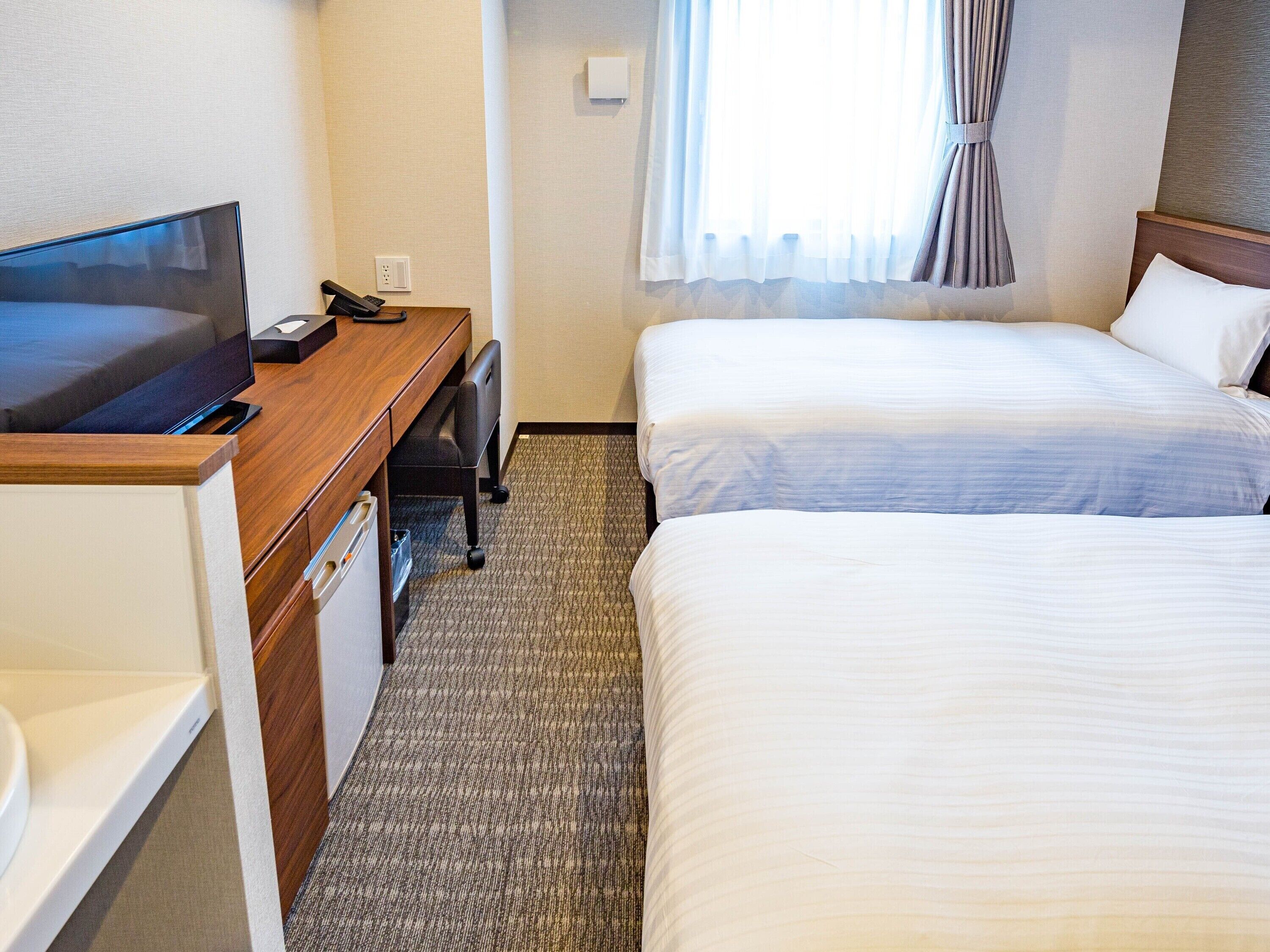 A hotel room with a desk, a television, two beds, and a window with curtains.