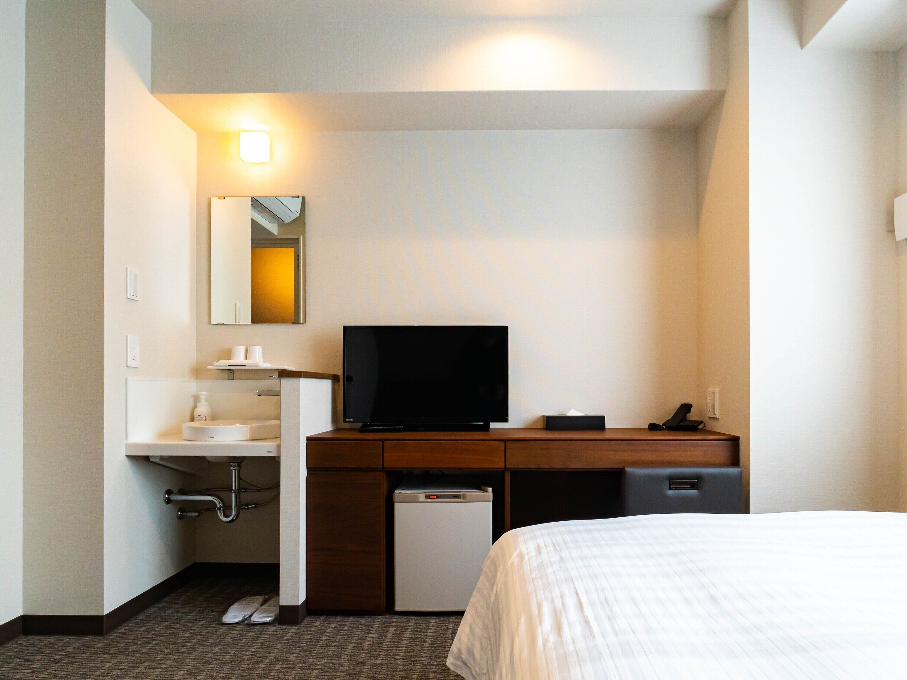 Ein Hotelzimmer mit einem Bett, einem Schreibtisch mit Fernseher, einer kleinen Kühlbox und einem Badezimmer mit Waschbecken und Spiegel.