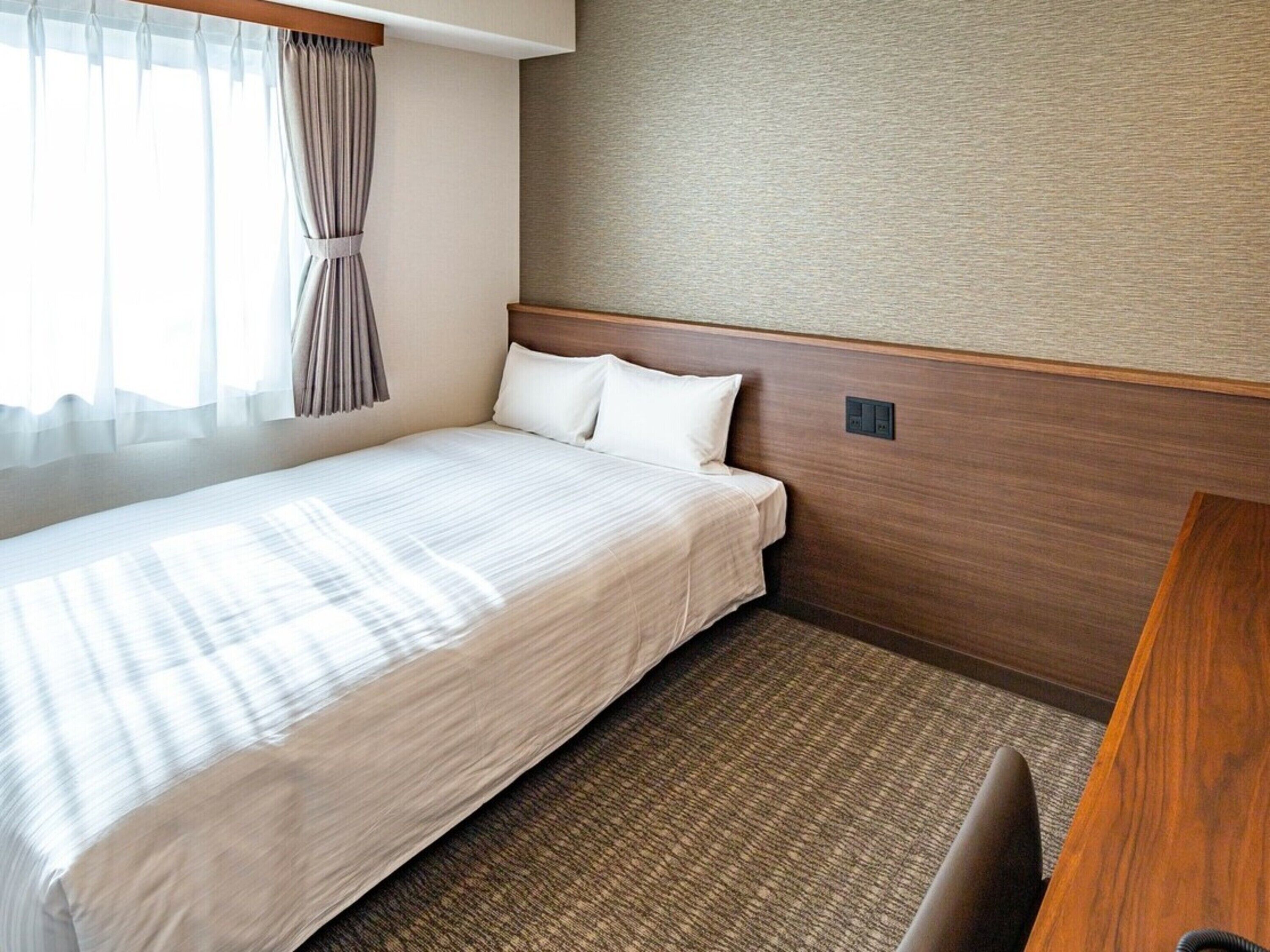 A hotel room with a bed, a wooden headboard, a carpeted floor, and a window with curtains.