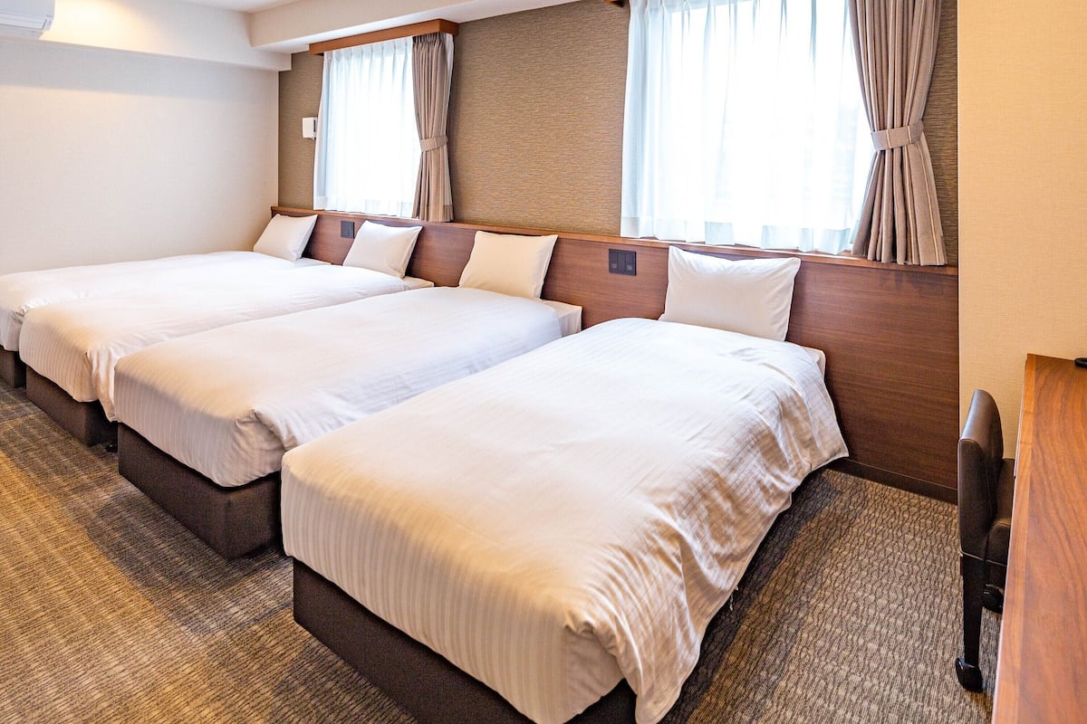 Habitación de hotel con tres camas individuales, cabecera de madera, suelo alfombrado y una ventana con cortinas.