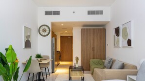 A modern hotel room with a bed, a sofa, a dining table, and a kitchen area visible through an open door.