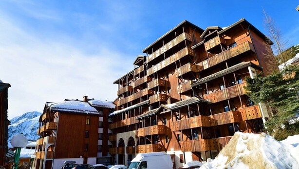 En flerlagsbygning i træ med balkoner, sandsynligvis et hotel eller resort, med sneklædte bjerge i baggrunden.