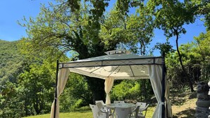 A covered outdoor dining area with a table and chairs, surrounded by trees and a stone wall.