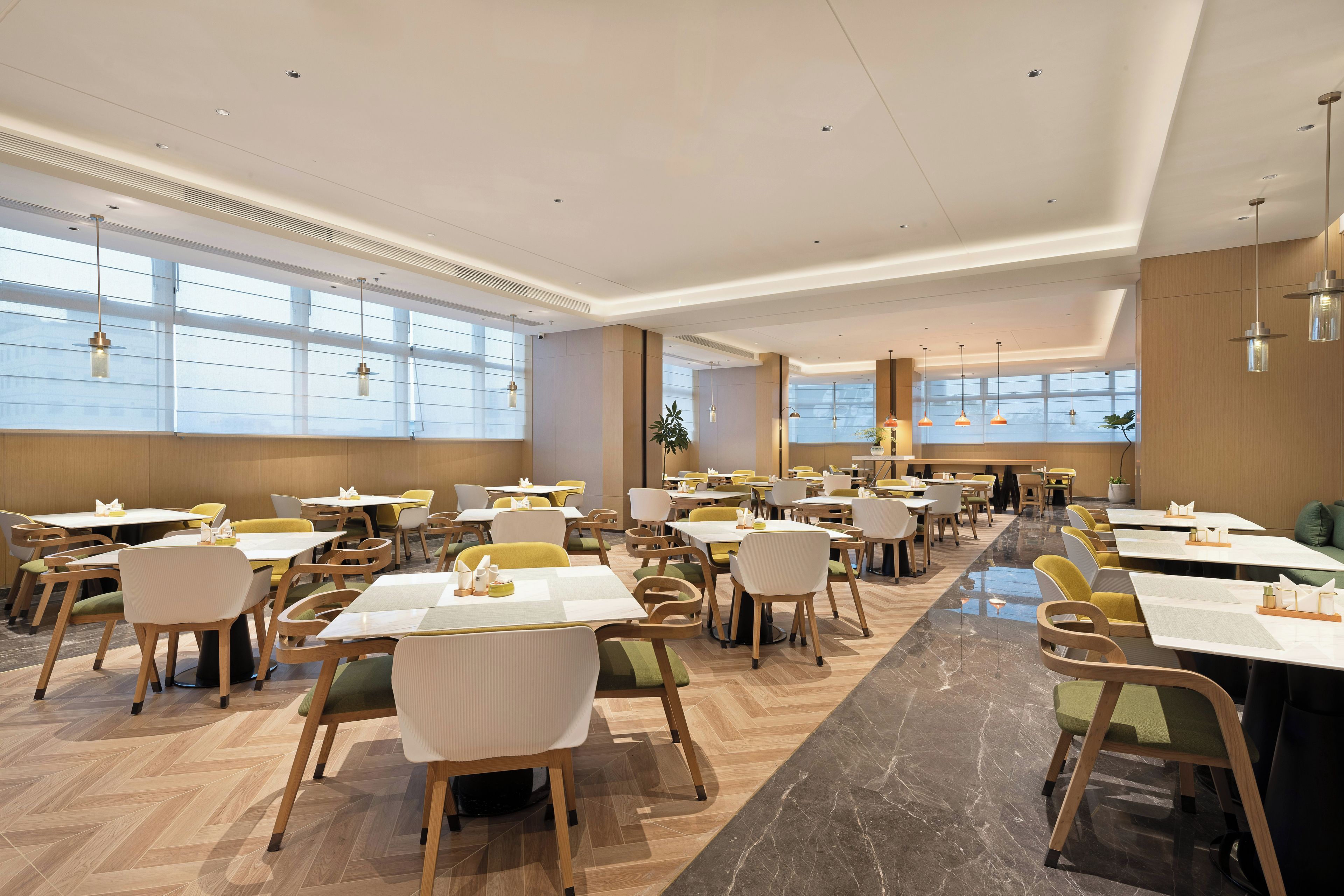 A spacious dining area with multiple tables and chairs, large windows, and a modern ceiling design.