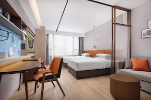 A modern hotel room with a bed, a desk, a chair, a sofa, and a small ottoman.