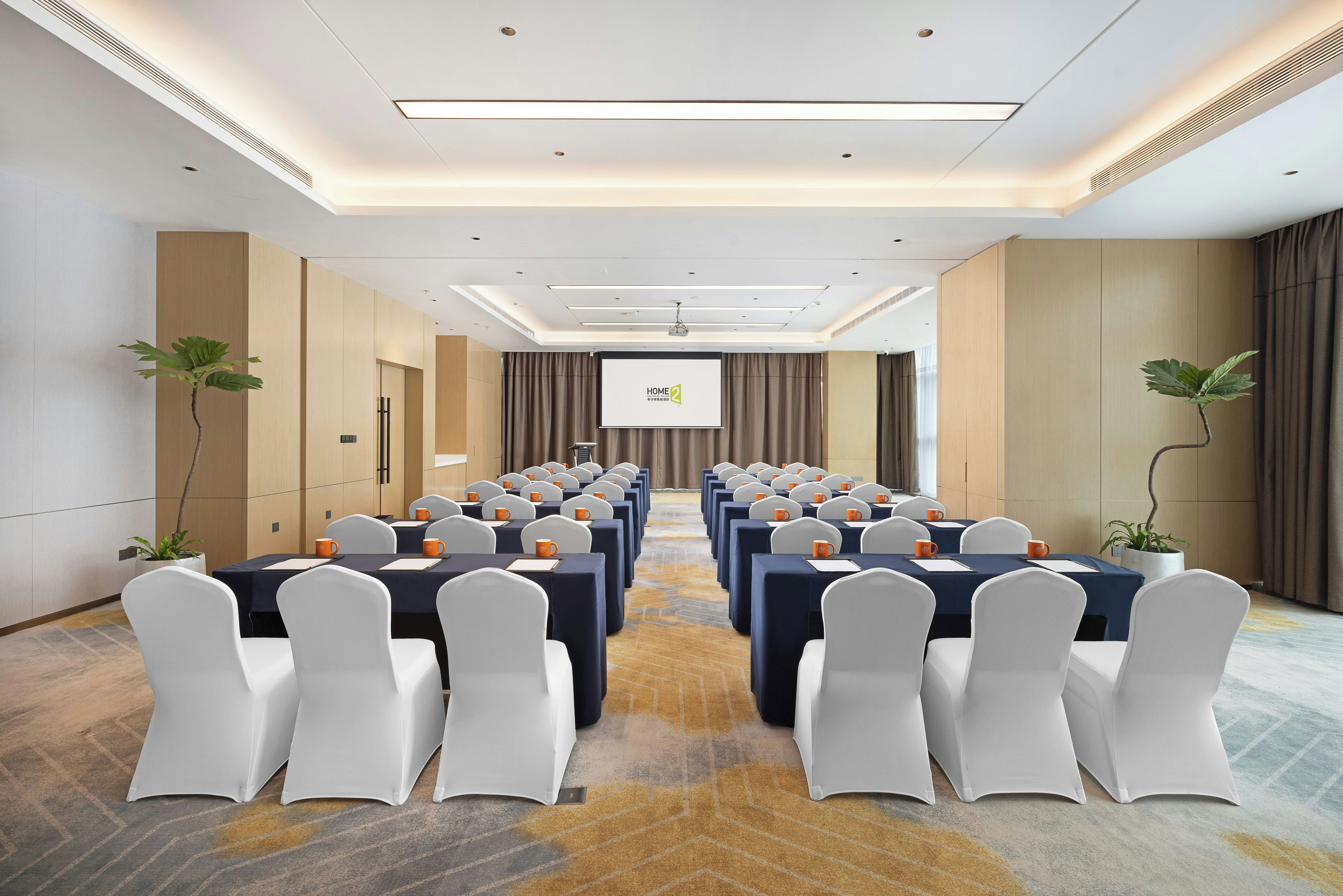 A conference room with rows of tables and chairs, a projector screen, and potted plants.