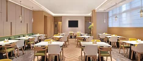 A hotel dining area with multiple tables and chairs, a flat-screen TV mounted on the wall, and a herringbone-patterned floor.