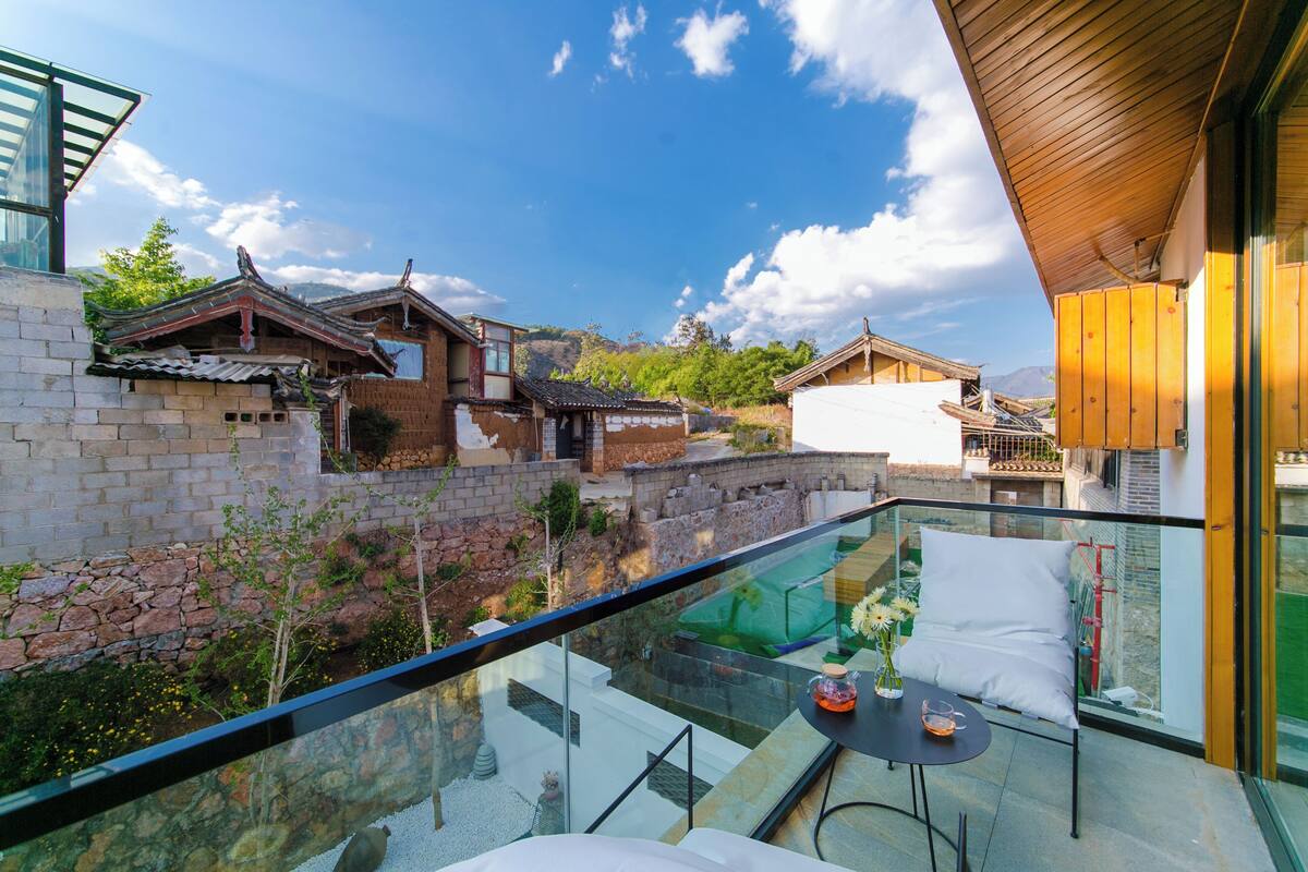 A balcony with a glass barrier, a table with a vase and a cushion, and traditional-style buildings in the background.