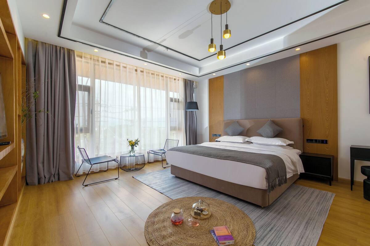 A modern hotel room with a large bed, a round coffee table, a desk, and a view of the city through large windows.