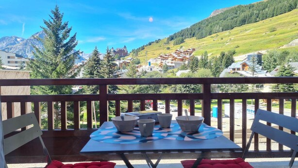 Un balcón con una mesa dispuesta para cuatro personas, con vista a un paisaje montañoso con edificios y follaje.