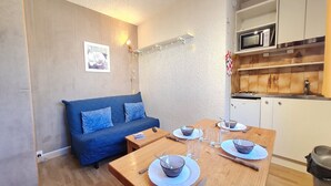 A compact living space with a blue sofa, a wooden dining table set with bowls and cutlery, a microwave, and a small kitchen area.