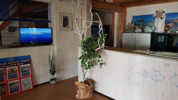 Un espace d’accueil doté d’un comptoir en bois, d’un téléviseur diffusant des images de paysages, d’un arbre décoratif et d’un mur orné d’œuvres d’art sur le thème maritime.