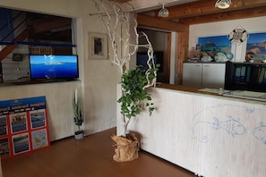 Un área de recepción con un mostrador de madera, un televisor que muestra una vista panorámica, una planta decorativa y una pared con arte temático marítimo.