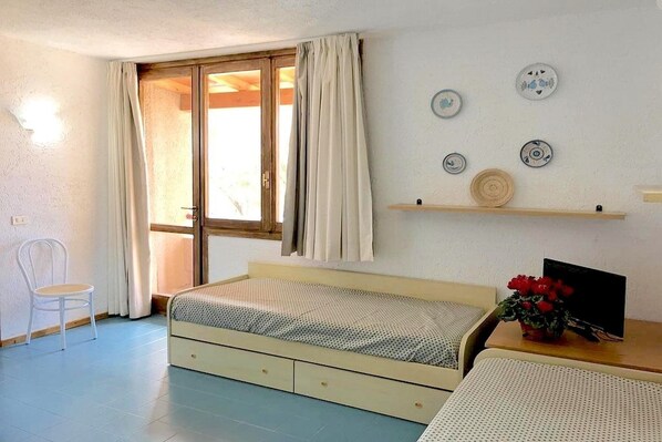 Un dormitorio con cama, una silla, un escritorio con computadora y una ventana con cortinas.