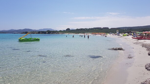 Ein Strand mit klarem Wasser, einer grünen Luftmatratze und Personen in der Ferne.