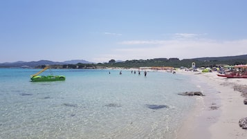 Ein Strand mit klarem Wasser, einer grünen Luftmatratze und Personen in der Ferne.
