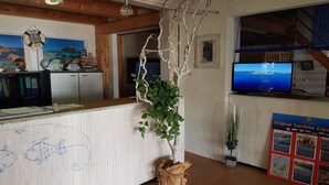 Ein Empfangsbereich mit einem hölzernen Empfangstresen, einem großen Bildschirm der eine Landschaft zeigt, einer an der Wand befestigten Rettungsboje und einem Plakat über Sardinien.