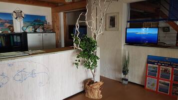 Ein Empfangsbereich mit einem hölzernen Empfangstresen, einem großen Bildschirm der eine Landschaft zeigt, einer an der Wand befestigten Rettungsboje und einem Plakat über Sardinien.