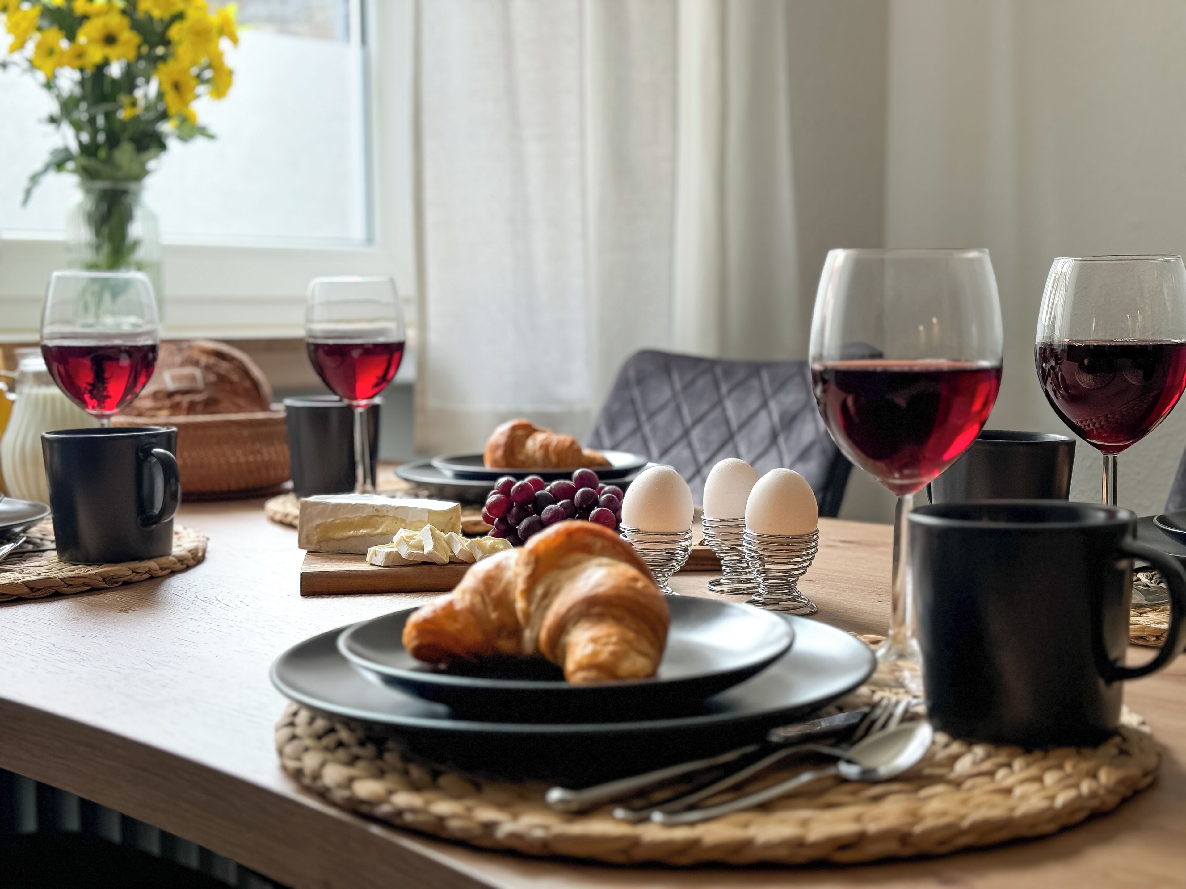 A breakfast setting with croissants, cheese, eggs, grapes, and wine glasses on a wooden table.