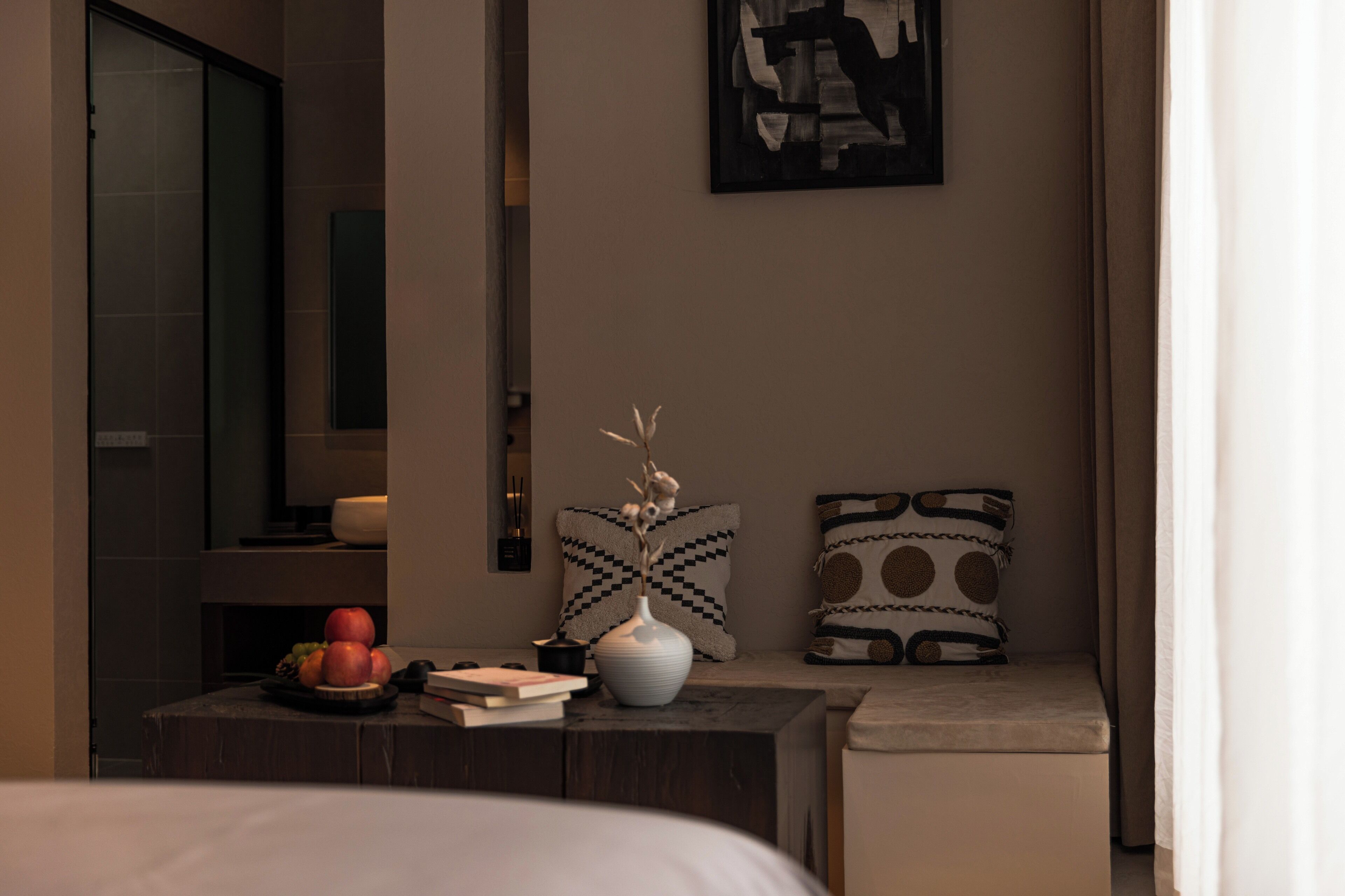 Habitació d'hotel moderna amb un llit, una taula de nit amb llibres i un vas, una taula petita amb fruita, i almoades estampades.