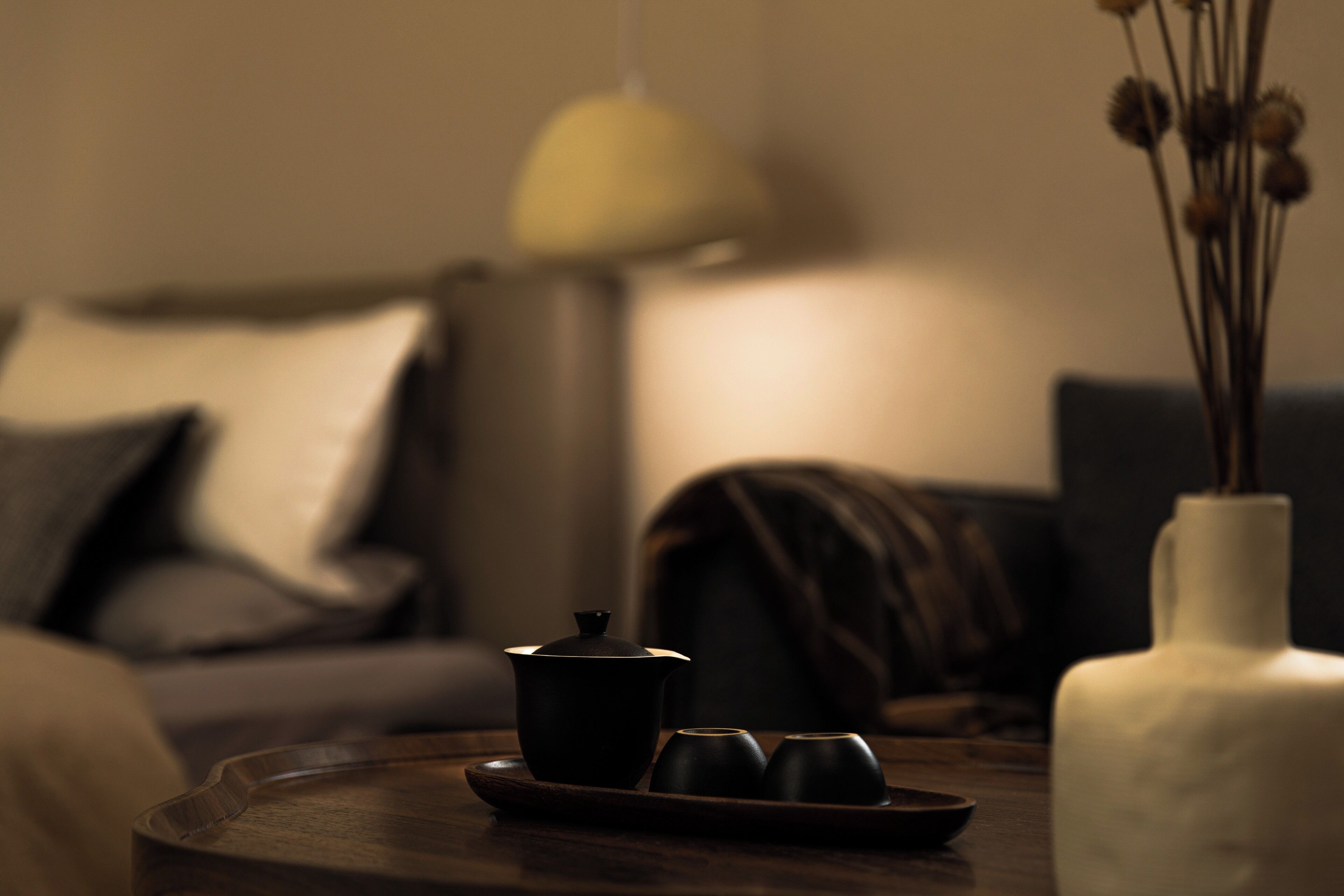 A cozy living room with a dark wooden coffee table, a black teapot, and two black cups. A white vase with dried flowers is on the right.