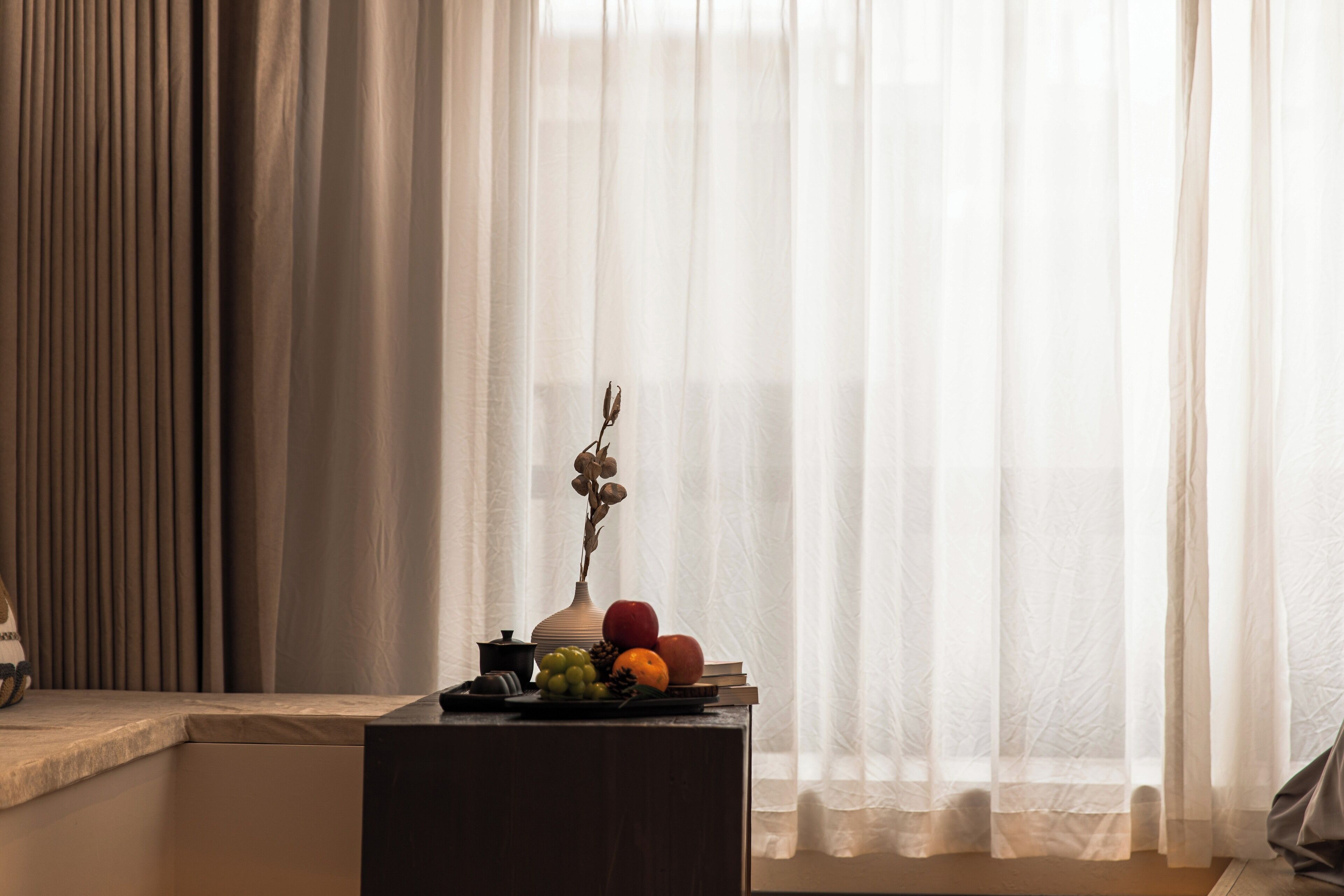 A room with a window covered by sheer curtains, a small table with a vase of dried flowers, and a fruit arrangement.