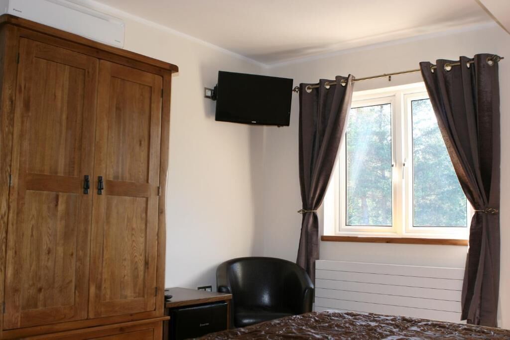 Quarto com guarda-roupa de madeira, uma poltrona de couro preto, cama, televisão de tela plana na parede e janela com cortinas.