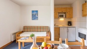 A small, cozy room with a bed, a table with fruits, a microwave, and a coffee maker in the kitchen area.