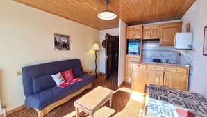 A compact living space with a sofa, a small table, a patterned bed, wooden cabinets, a tiled backsplash, and a mounted television. - Ski-In Apartment with Balcony (Les Deux Alpes)