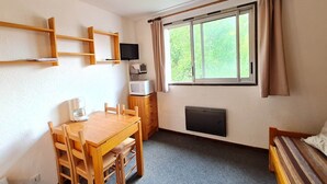 Una habitación pequeña con una mesa de madera, sillas, cama, televisión, calefactor y ventana con cortinas.
