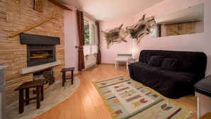 A living room with a stone fireplace, a black sofa, a patterned rug, and a large wall hanging. - Studio in France with Mountain Views (Allos)
