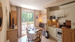 Una habitación acogedora con una mesa de comedor lista para cuatro personas, un sofá, una cocina con armarios y electrodomésticos blancos, y un ventanal con cortinas.
